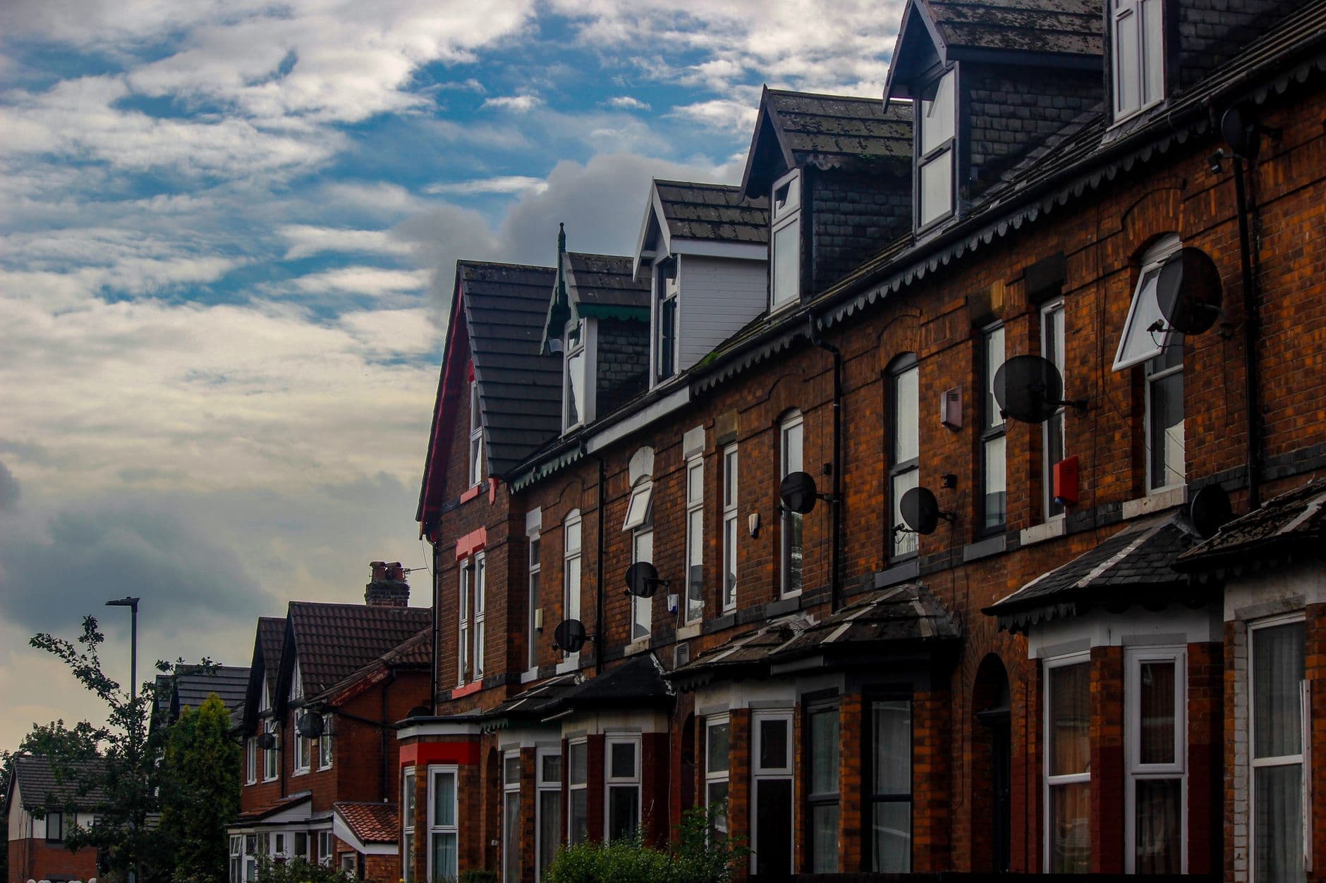 A row of red-brick Victorian terraced houses in Manchester, satellite dishes on the fronts, under a partly cloudy sky.