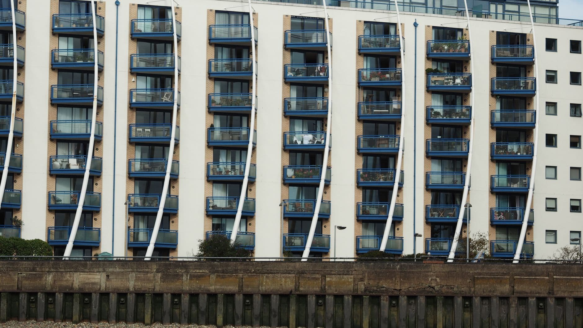 A riverside block of modern flats with rows of identical blue balconies above a river wall.