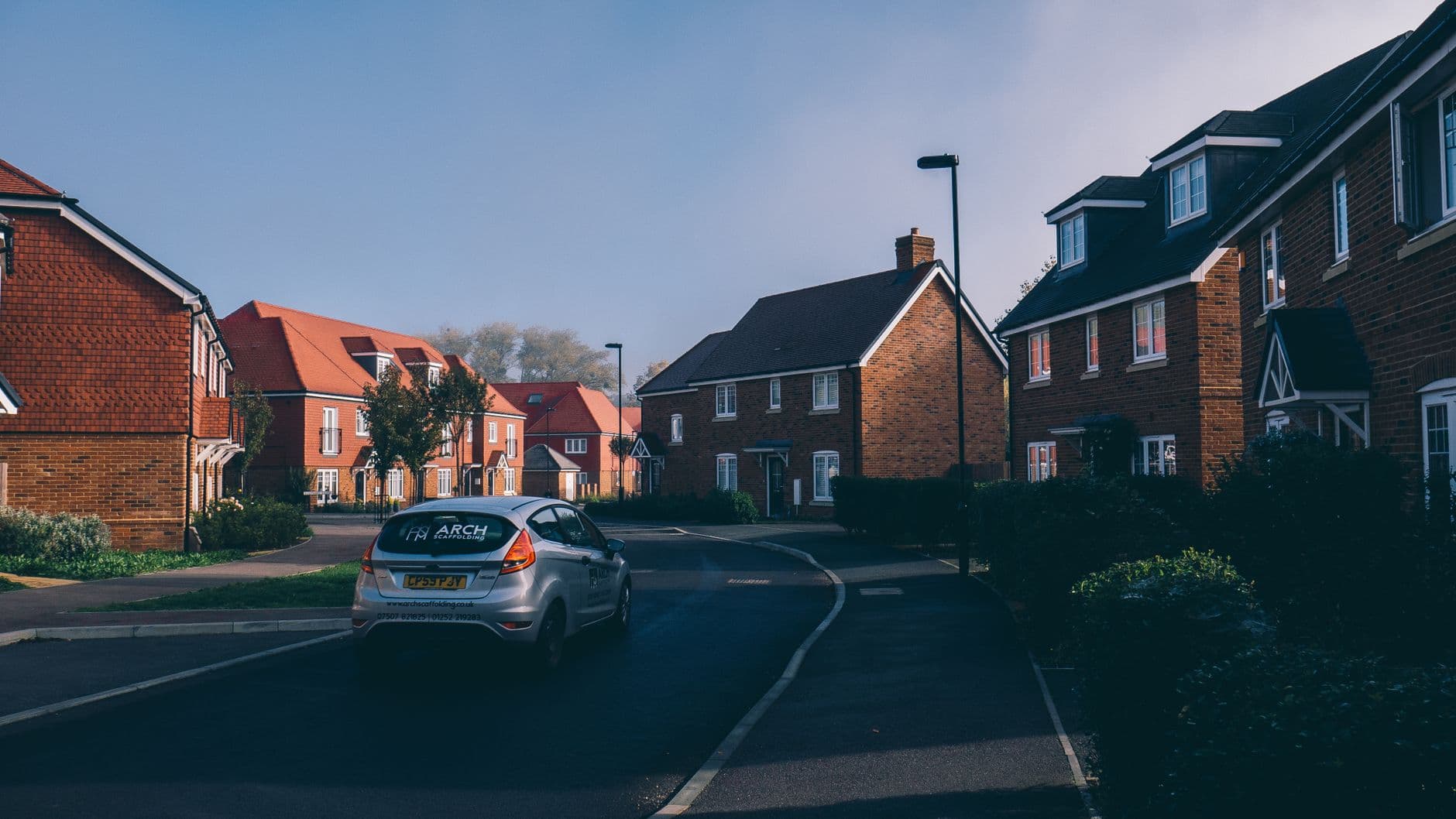A curving street of recently built brick houses on an English estate, with a car driving through in soft morning light.