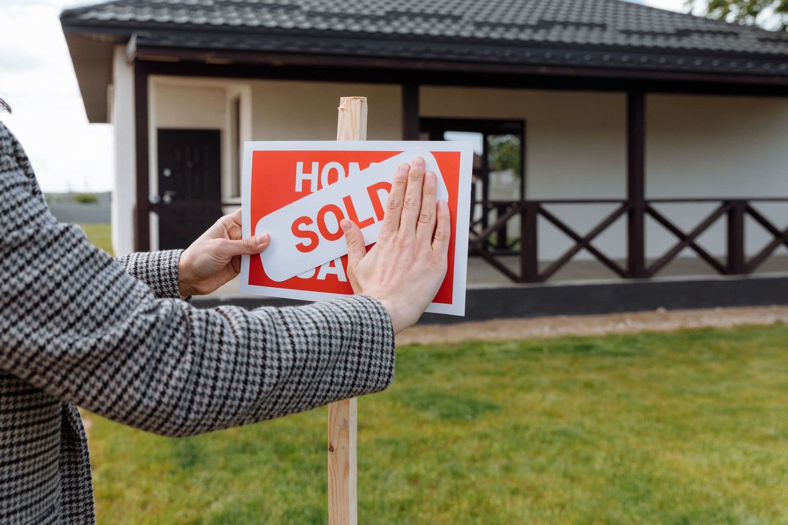 Hands placing a 'sold' sticker on a for-sale sign in front of a house